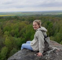 Auf dem Foto ist Wibke Sophie Meurer sitzend und in die Kamera über ihren Rücken schauend auf einem Feld mit Blick über einen Wald zu sehen.
