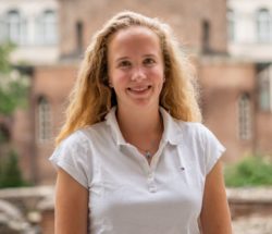 Auf dem Foto ist Ronja Horacek lächelnd und im Hintergrund eine Reihe Backsteinhäuser zu sehen.