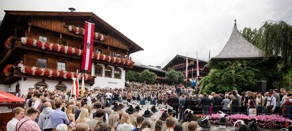 25-08-26_Eröffnung EFA_Banner Auf dem Foto sieht man eine Menschenmenge in traditioneller österreichischer Kleidung, die einemStraßenumzug anlässlich der Eröffnung des European Forum Alpbach beiwohnt.