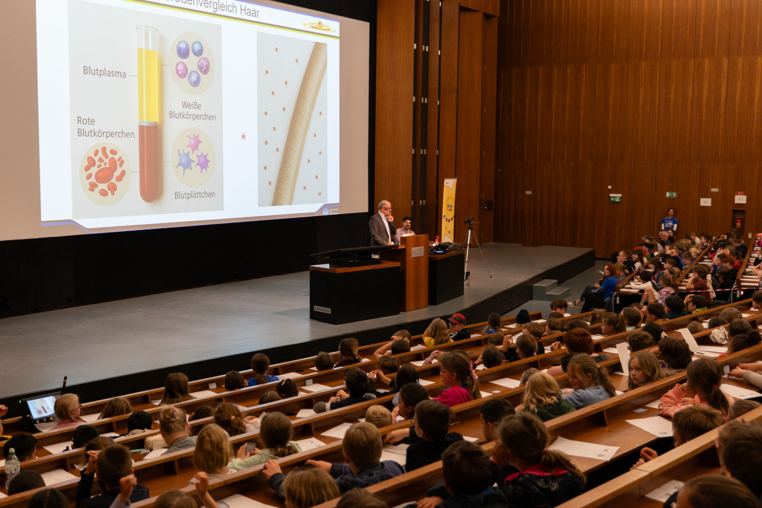 Sur la photo, on voit Christian Wagner, responsable de programme, donner une conférence à la Kinderuni sur le thème du sang. Au premier plan, on aperçoit son jeune public vu de dos.